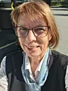 Nærportrett av Aase Marie Anthonisen med briller, vest og nøkkelbånd ved en buss.