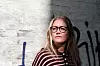 Kristine Wennerberg med briller og stripet genser foran en murvegg med graffiti.