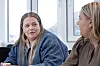 To kvinner sitter i samtale ved et vindu, iført denimjakke og brun kjole.