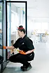 Astri Karine Lundgren huker ved en åpen kjøledisk i kantinen, iført svart uniform og oransje hansker.