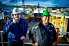 To arbeidere med vernehjelm står på et industrielt anlegg med tørrdokk og bebyggelse i bakgrunnen.