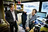 To menn står og sitter på et kontor med datamaskiner, kontorstoler og hyller med permer.
