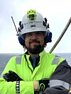 Arbeider i grønn synlighetsjakke og verneutstyr står foran havet.