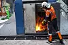 Arbeider i oransje verneklær står foran en åpen industriovn med synlig flamme.