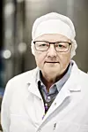 Fabrikksjef Roar Moss med briller, hårnett og hvit laboratoriefrakk foran en uskarp bakgrunn.