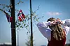 Kvinne i bunad foran norske flagg og blå himmel.