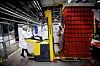 To arbeidere håndterer varer ved en palletruck i en fabrikk, med en høy stabel røde kasser til høyre.