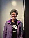 Alexandra Coelho smiler foran en lys vegg, iført lilla jakke, briller og svart T-skjorte.