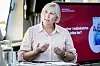 Anne Berit Aker Hansen snakker ved et bord foran en rød presentasjonsskjerm.
