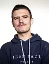 Portrett av Jon Philip Bjerklund i mørk hettegenser med Jean Paul-logo mot lys bakgrunn.