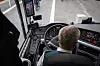 Bussjåfør sett bakfra ved rattet i en moderne buss med instrumentpanel og skjermer.