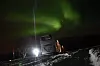 Grønt nordlys over en snødekt vei ved Skarsfjord, med et kjøretøy og lysende frontlykter.