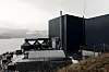 Industrianlegg fra Nordic Mining ved fjorden på Engebø, med mørke bygninger og fjell i bakgrunnen.